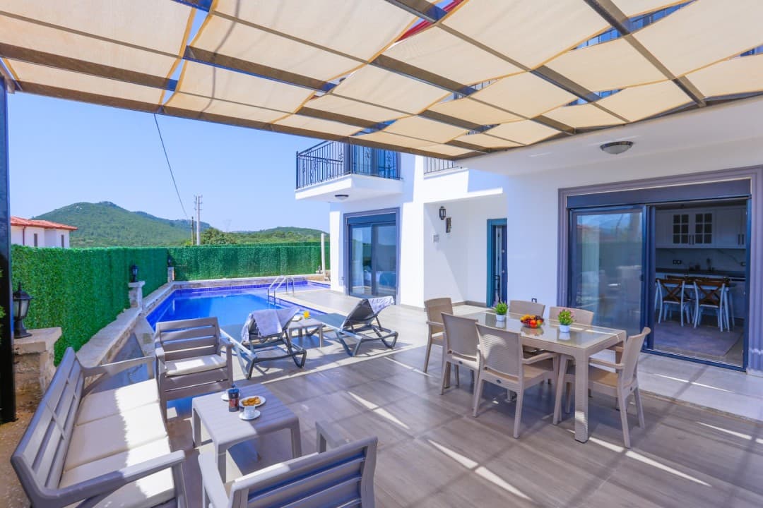 Güneşlikli terasta yemek masası, oturma grubu ve yanında açık hava yüzme havuzu bulunan beyaz villa.