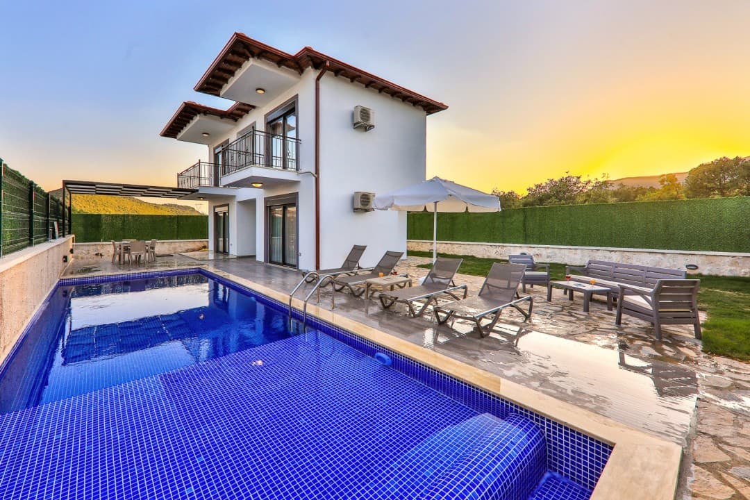 Gün batımında havuz başında şezlonglar, güneş şemsiyesi ve oturma grubu bulunan iki katlı beyaz villa.