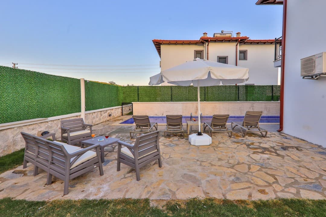 Taş zeminli terasta oturma grubu, şemsiye ve arka planda havuz.