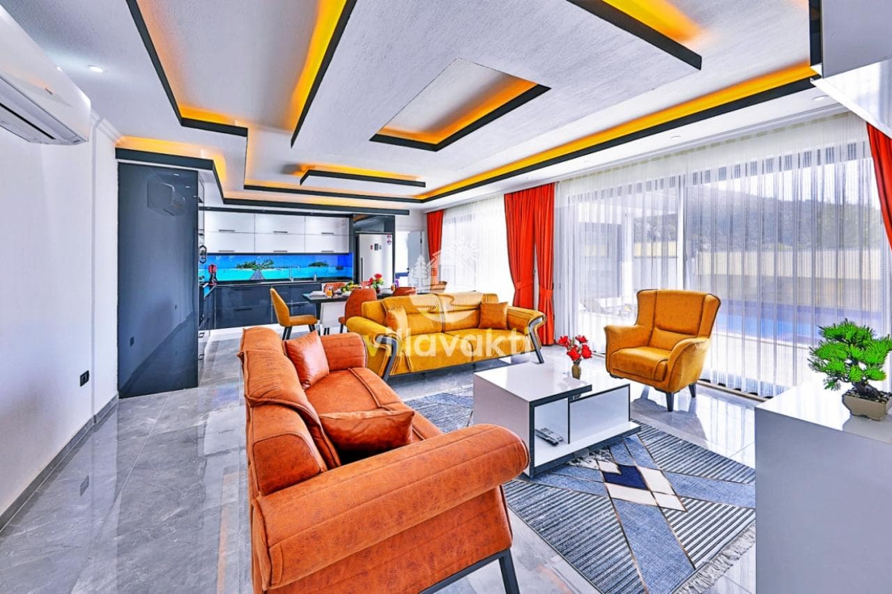 Modern salon, turuncu koltuklar, beyaz mutfak dolapları ve asma tavan aydınlatması.