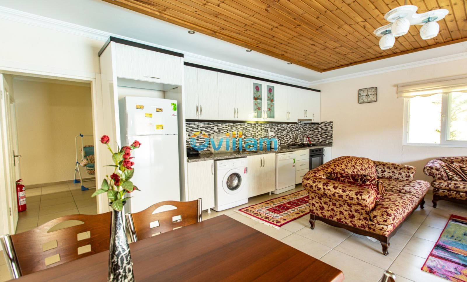 Ahşap tavanlı, desenli koltuk takımı, beyaz mutfak dolapları ve yemek masası içeren açık mutfaklı salon.