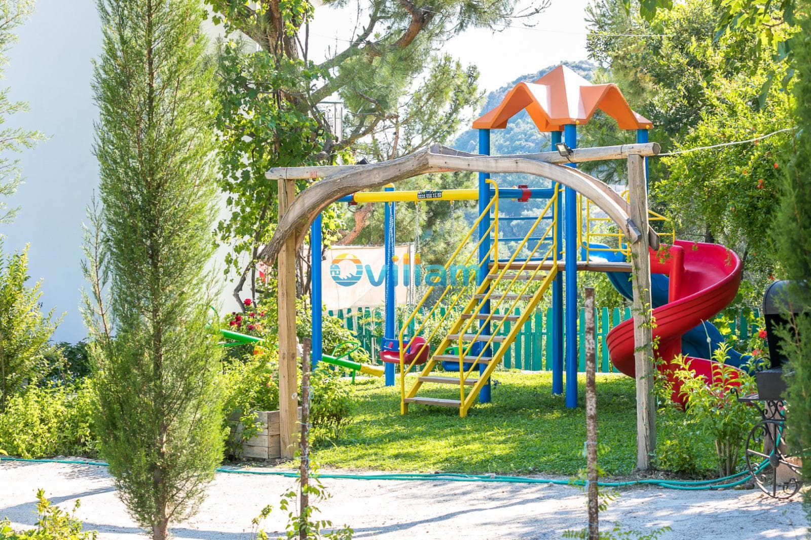 Yeşil bahçe içinde renkli çocuk oyun parkı, kaydırak ve salıncak.