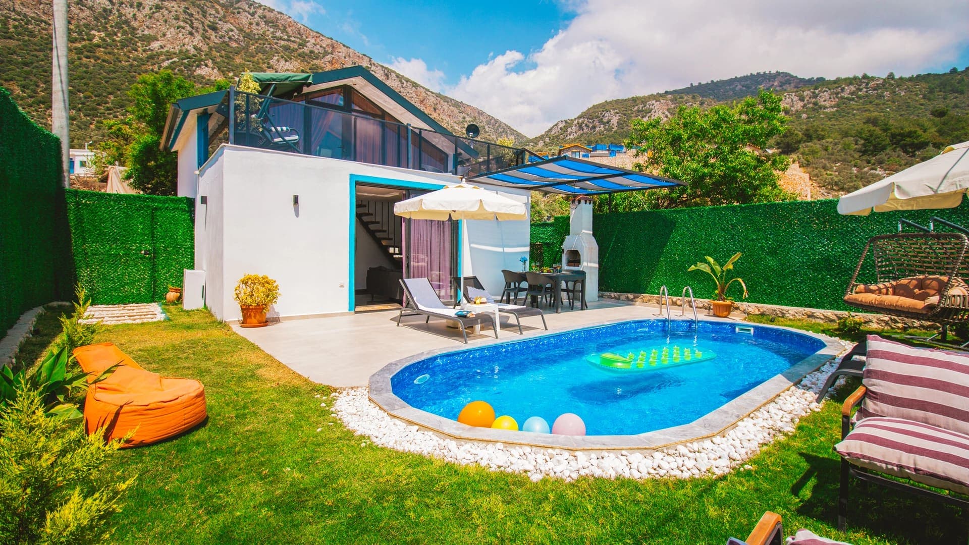 Yeşil çitlerle çevrili bahçesinde havuz, şezlonglar, salıncak ve dış mekan yemek masası olan villa.