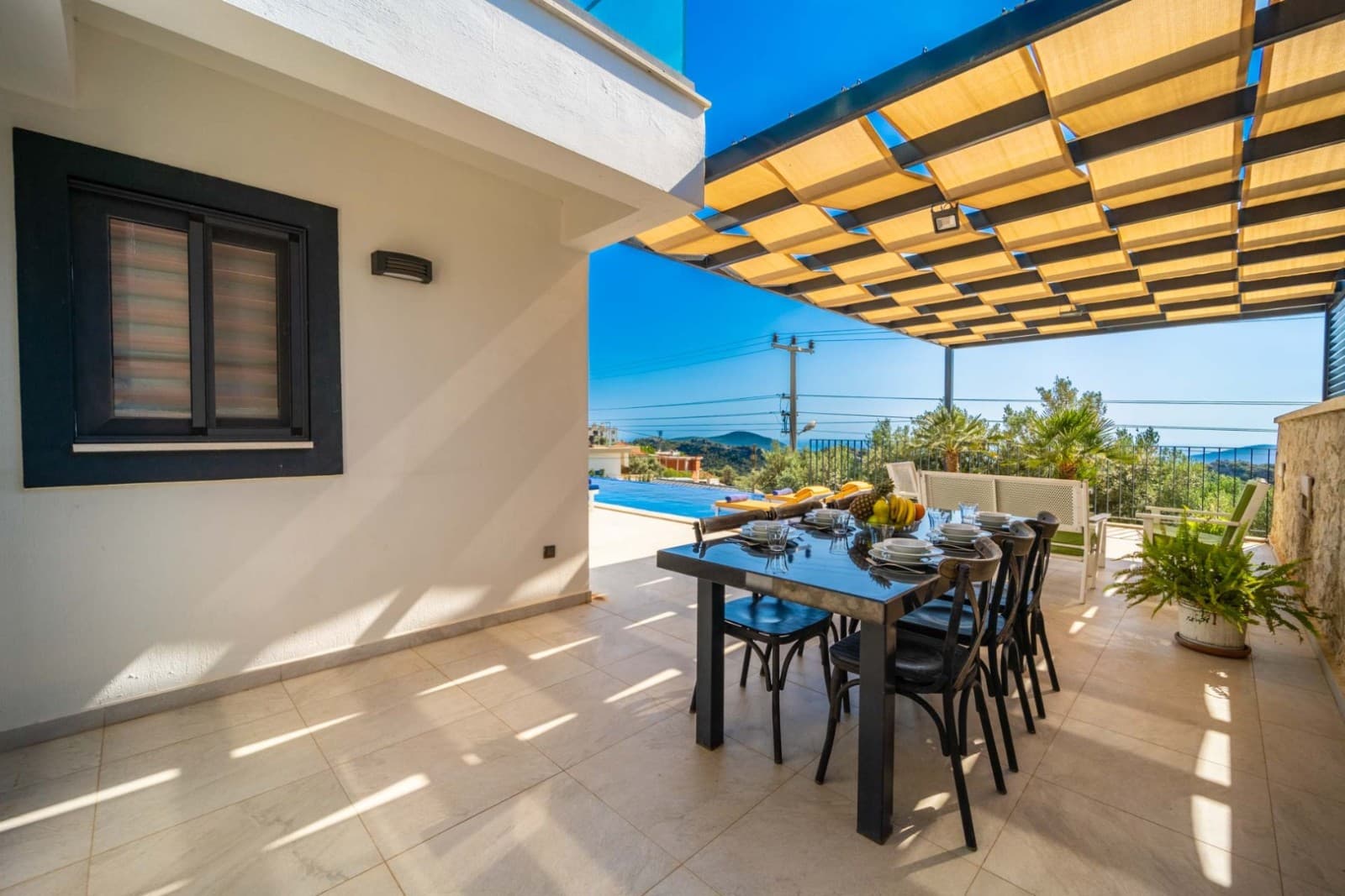 Güneşlikli terasta yemek masası ve sandalyeleri, arka planda deniz ve doğa manzarası.