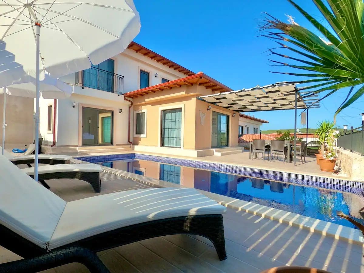 Havuz başında beyaz şezlonglar, güneş şemsiyesi ve gölgelikli yemek masası bulunan villa.