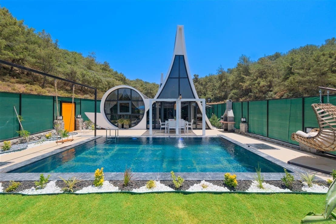 Önünde yüzme havuzu, şezlonglar ve çim alanı bulunan modern mimarili villa dış cephesi.