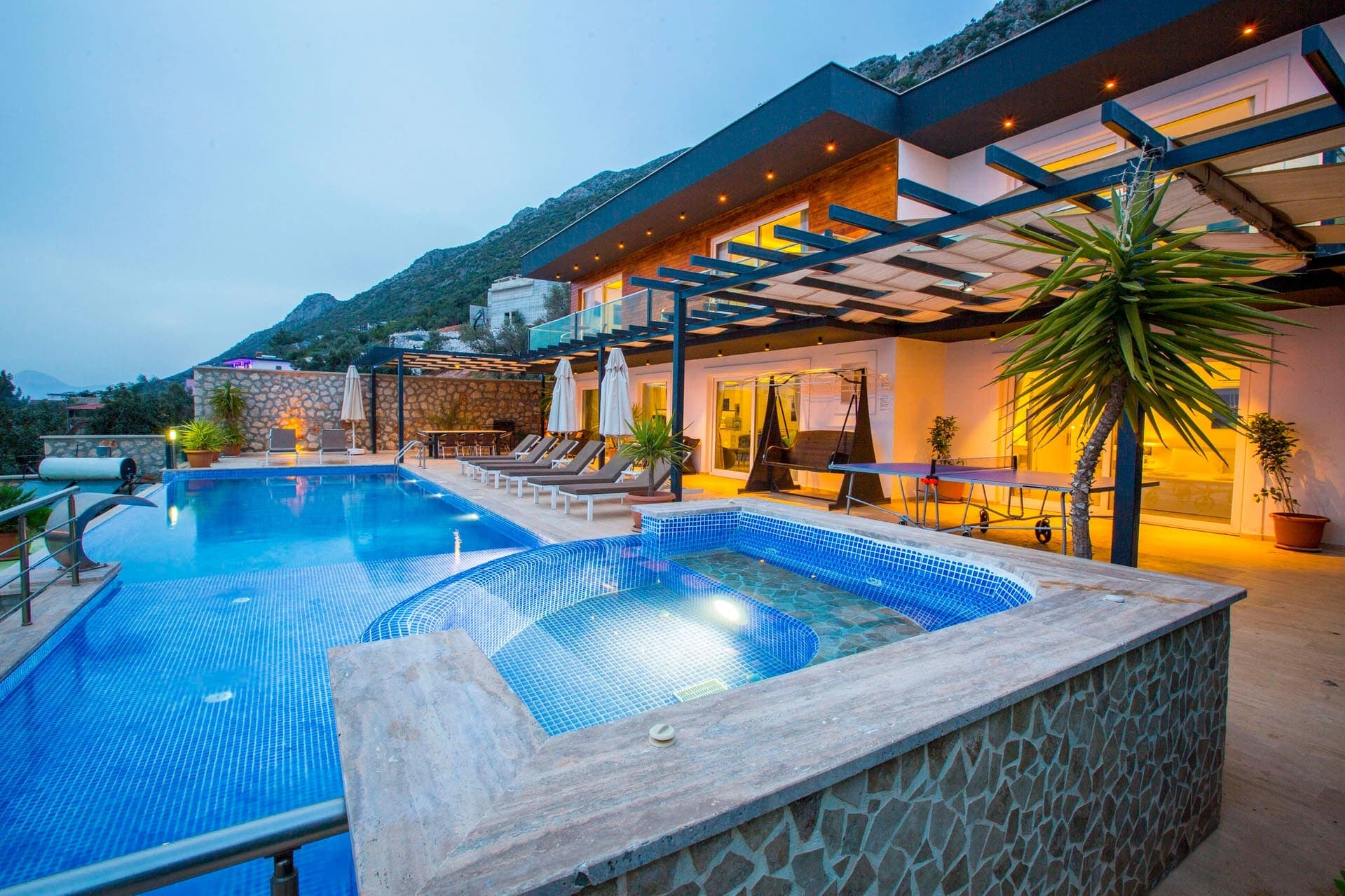 Akşam ışıklandırmalı havuz, jakuzi, şezlonglar ve bahçede masa tenisi bulunan modern villa dış cephesi.