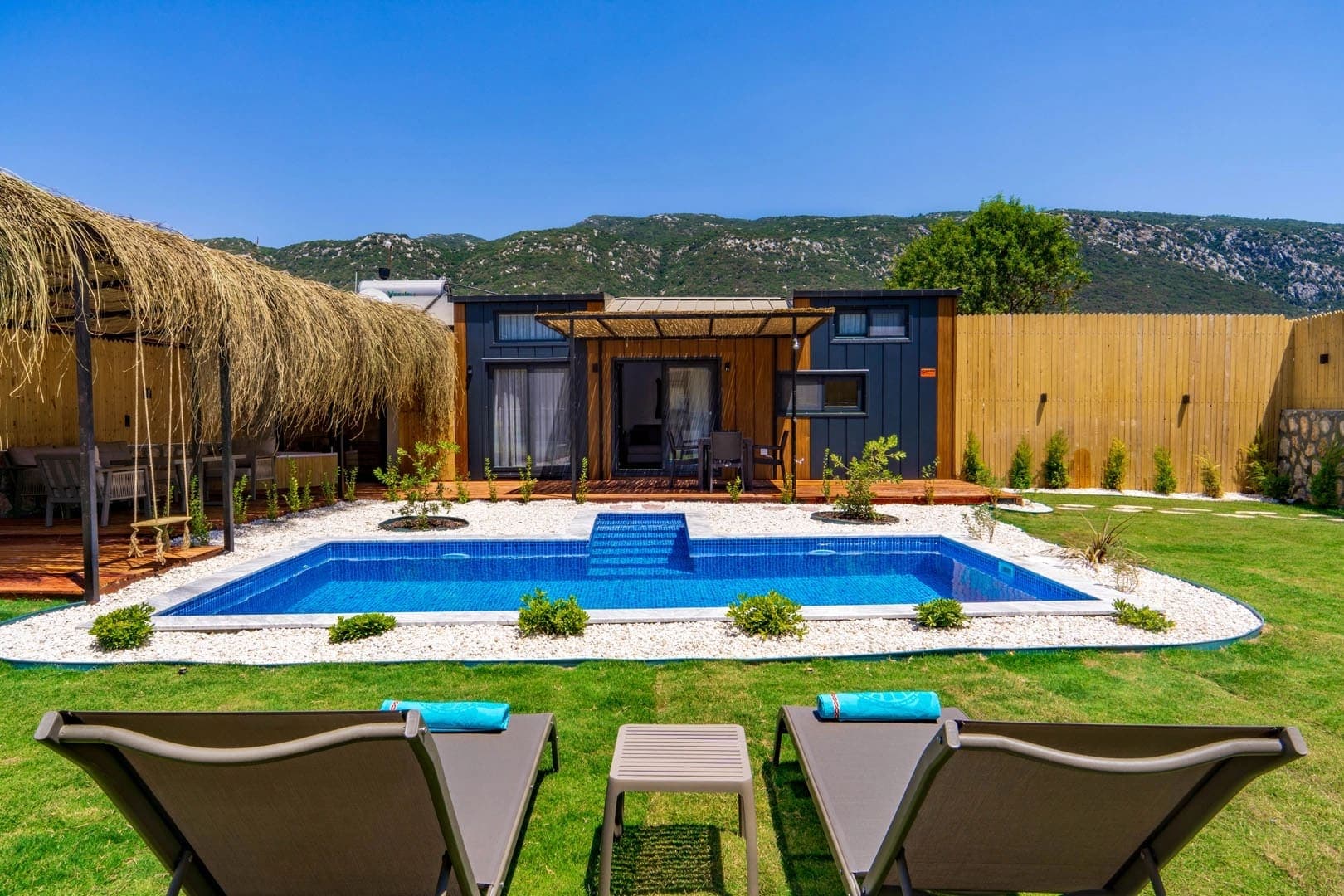 Önünde havuz ve şezlonglar bulunan, ahşap kaplamalı tek katlı küçük villa ve bahçesi.