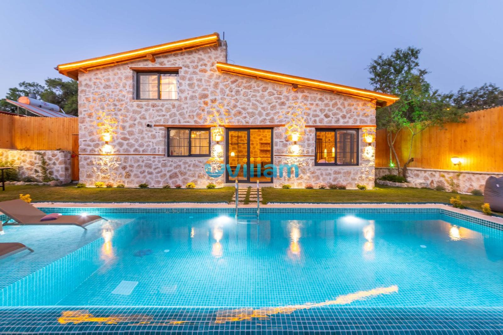 Akşam vakti aydınlatılmış taş villa ve önünde yer alan dikdörtgen yüzme havuzu.