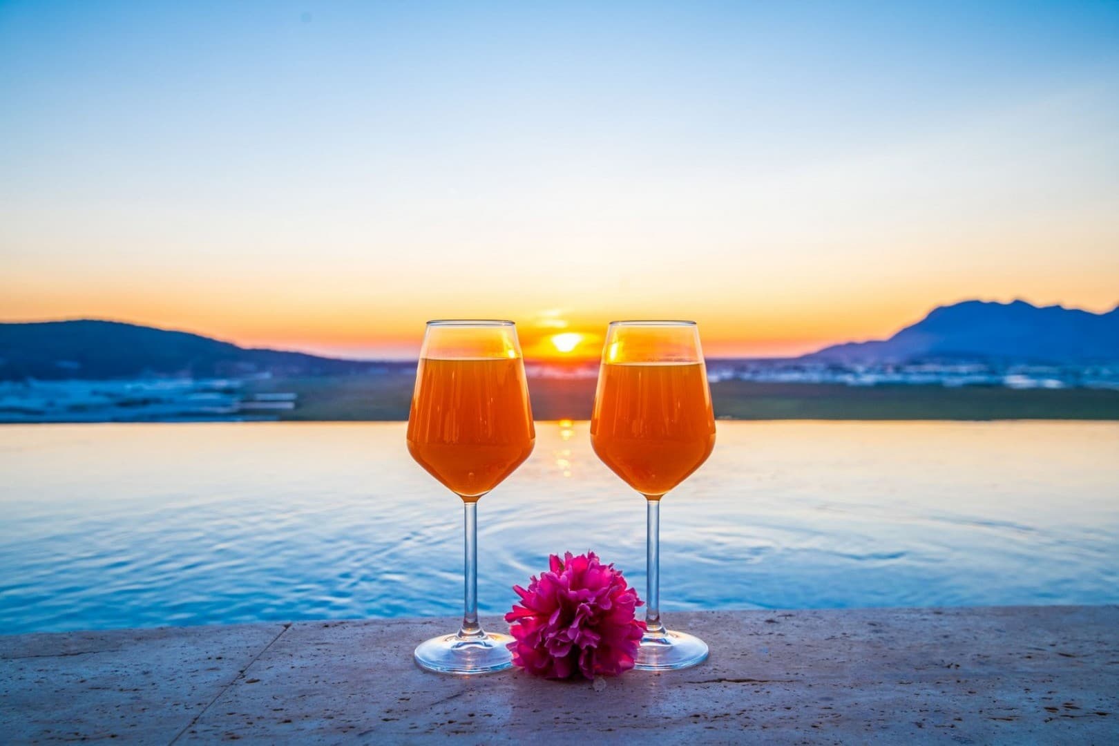 Havuz kenarında gün batımı manzarasına bakan iki adet kadeh ve pembe çiçek.