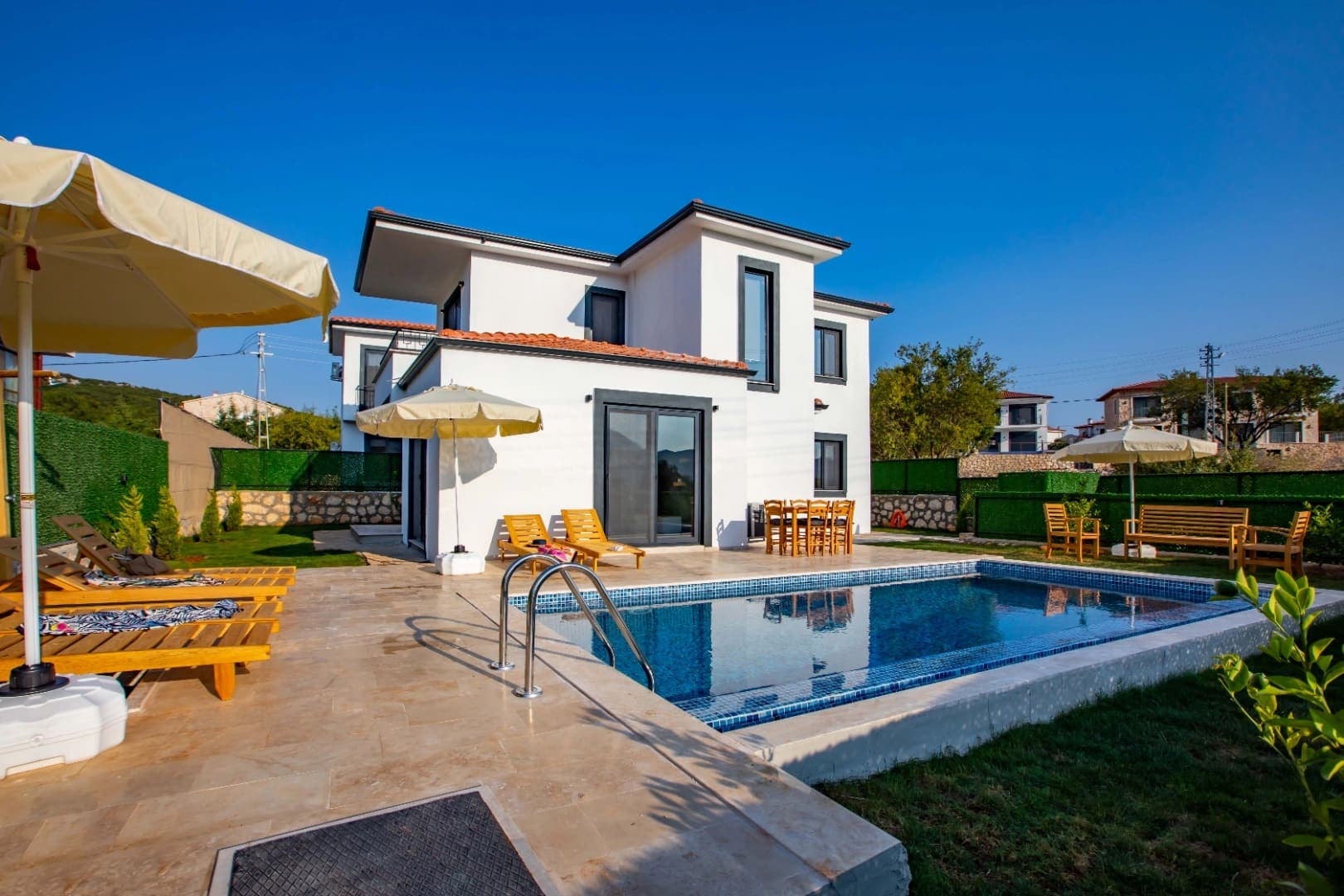 Beyaz dış cepheli villa, önünde güneş şemsiyeleri, şezlonglar ve mavi yüzme havuzu.