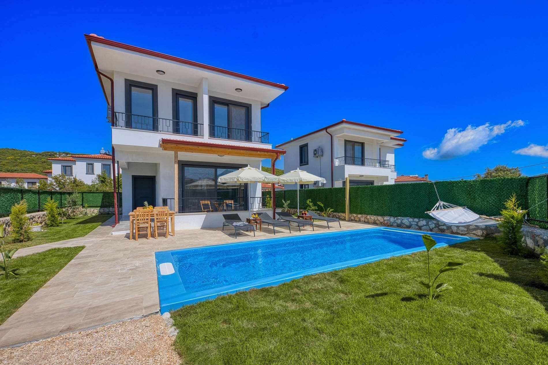 İki katlı beyaz villa, bahçesinde havuz, şezlonglar, yemek masası ve hamak.