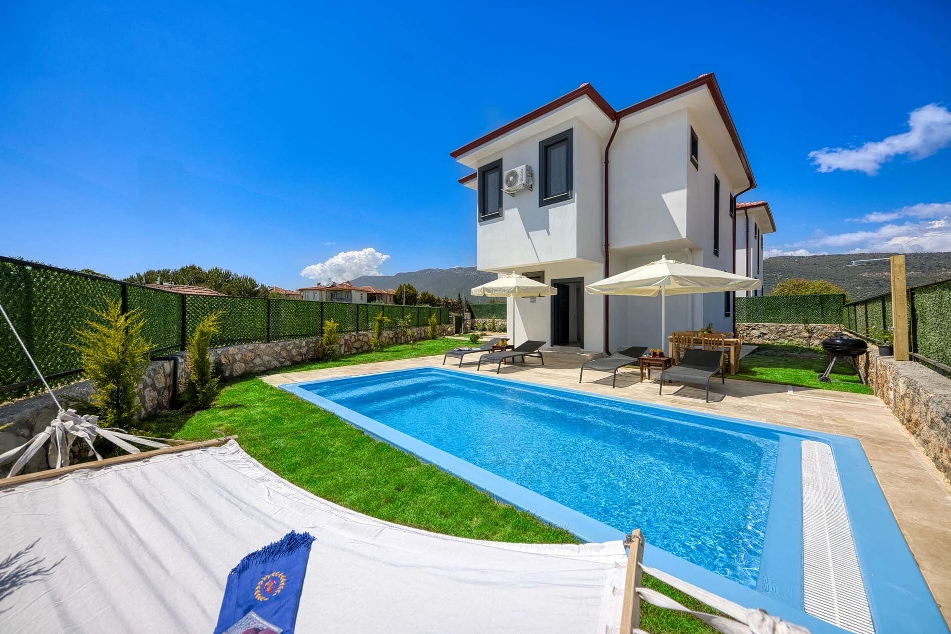 Mavi yüzme havuzu, şezlonglar, bahçe alanı ve arka planda beyaz villa.