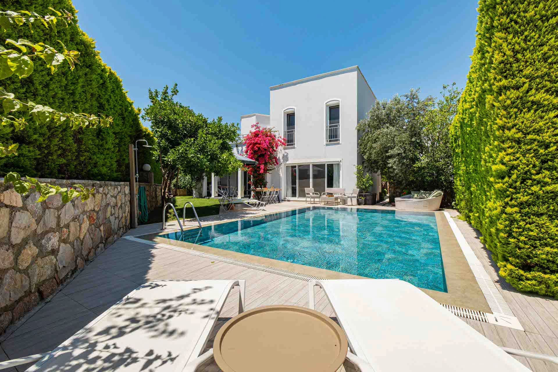 Yüksek çitlerle çevrili bahçede havuz, şezlonglar ve çiçekli beyaz villa.