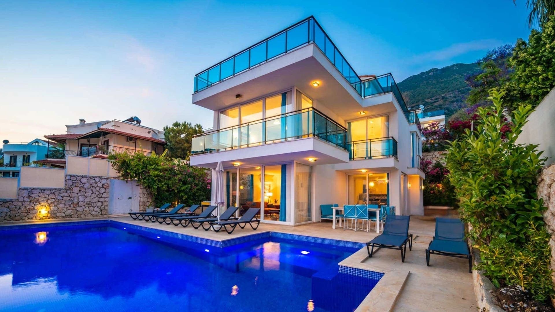 İki katlı modern villa, önünde şezlonglar bulunan havuz ve akşam aydınlatması.