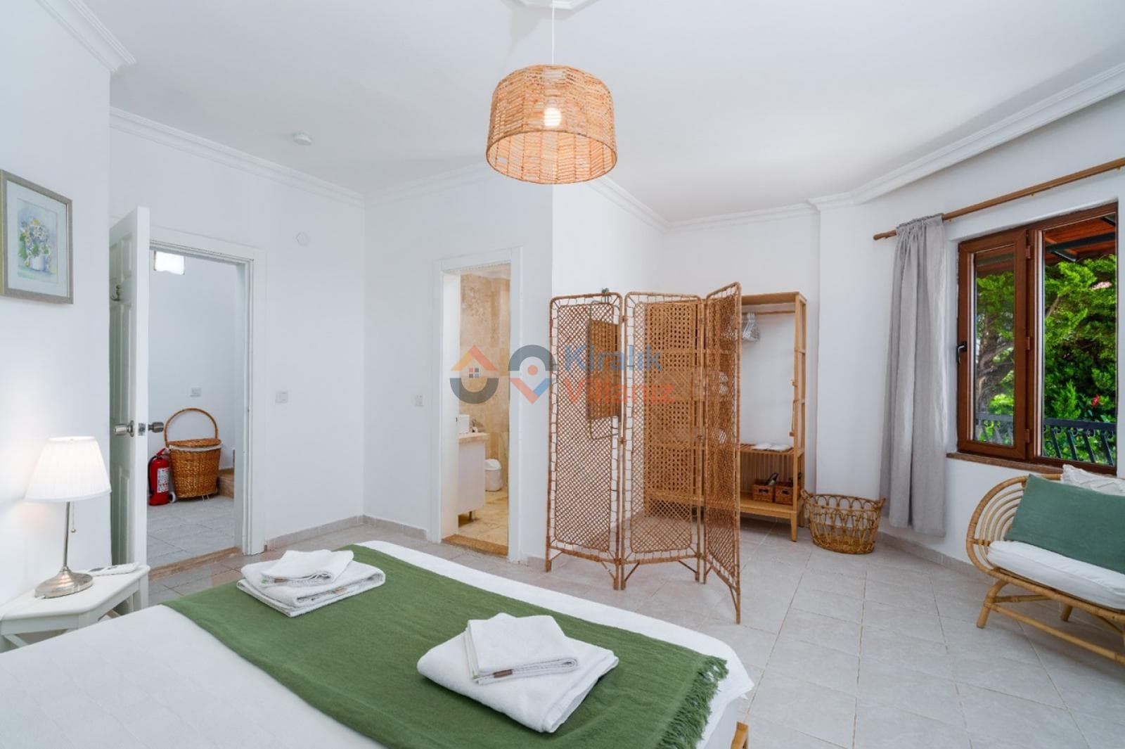 Yeşil örtülü yatak, hasır paravan, rattan koltuk ve banyo kapısı bulunan yatak odası.