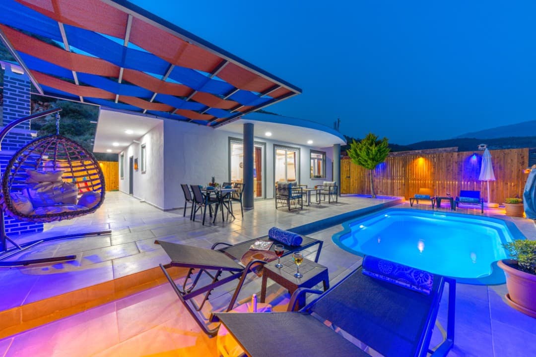 Akşam aydınlatmalı havuz kenarında şezlonglar, salıncak ve yemek masası bulunan villa terası.