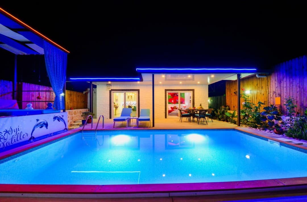 Mavi ışıklandırmalı açık havuz kenarında şezlonglar ve gece aydınlatılmış villa.
