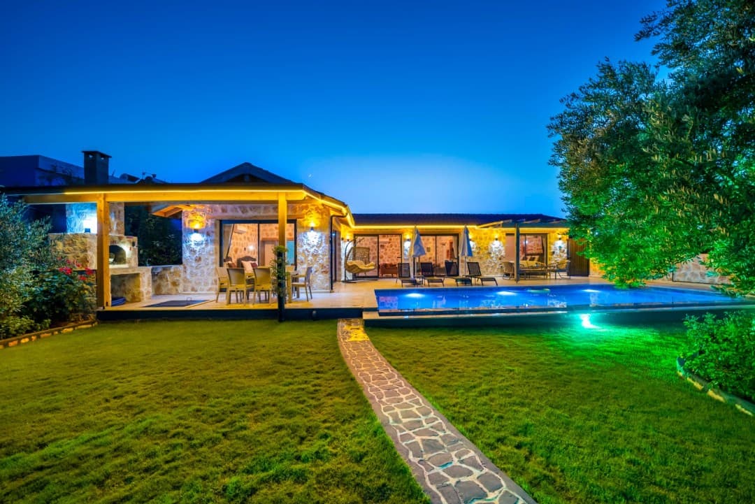 Akşam karanlığında sarı ışıklarla aydınlatılmış taş villa, havuz ve çim bahçe.