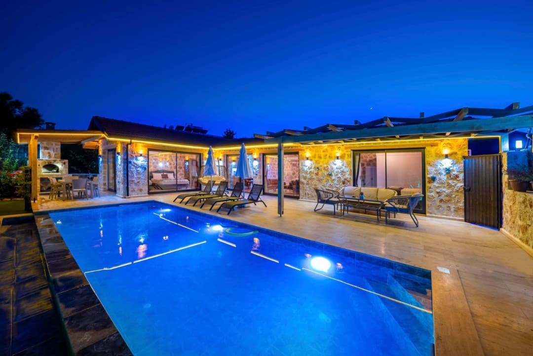 Gece ışıklandırılmış taş villa cephesi, özel havuz ve oturma grubu.