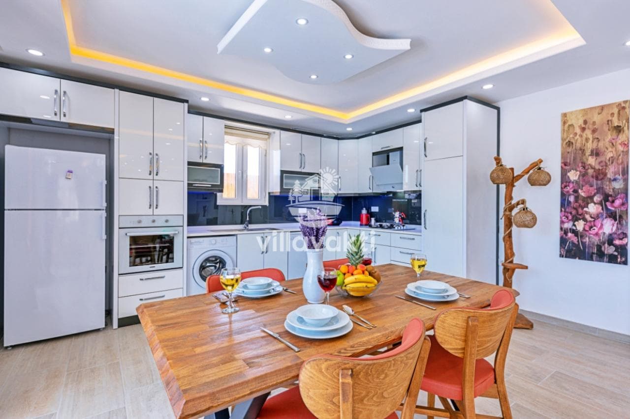 Ahşap yemek masası, beyaz mutfak dolapları ve modern aydınlatmalı mutfak alanı.