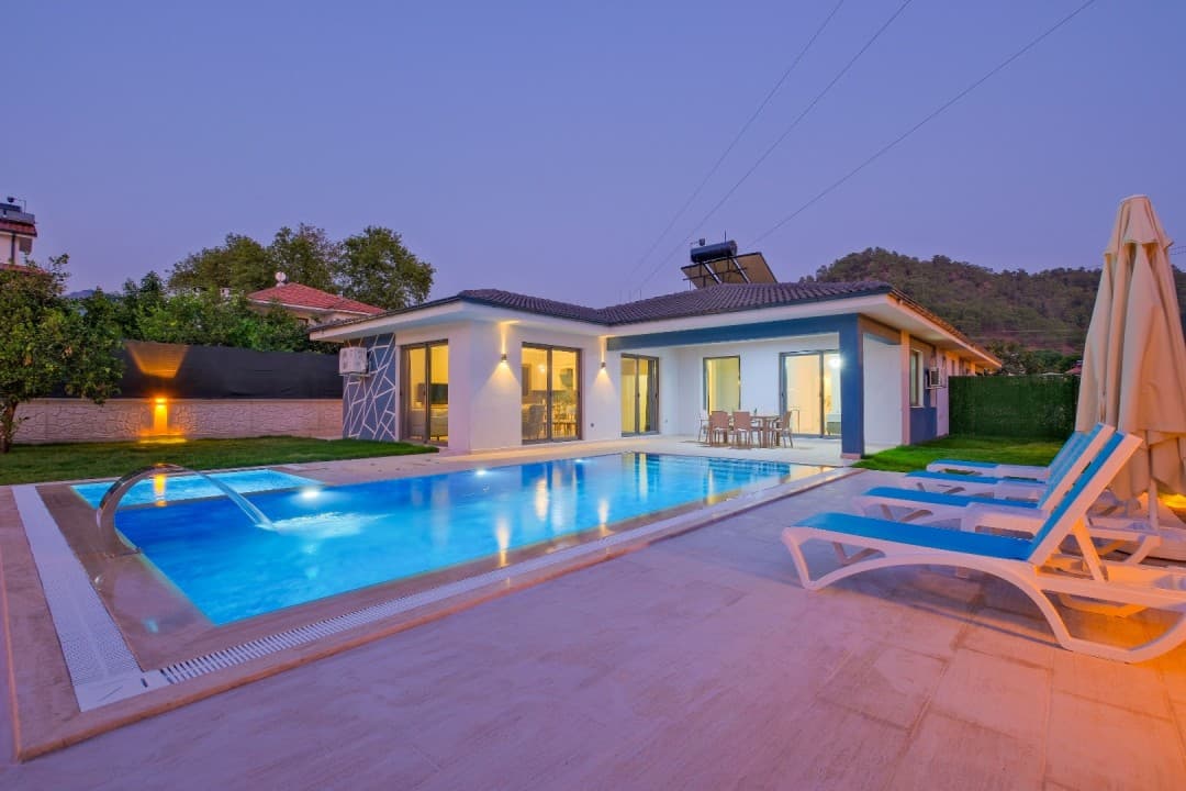 Aydınlatmalı yüzme havuzu ve şezlongların bulunduğu beyaz dış cepheli villa.
