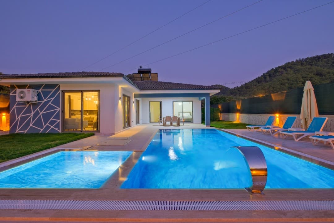 Akşam vakti aydınlatılmış havuz, şezlonglar ve tek katlı villa cephesi.