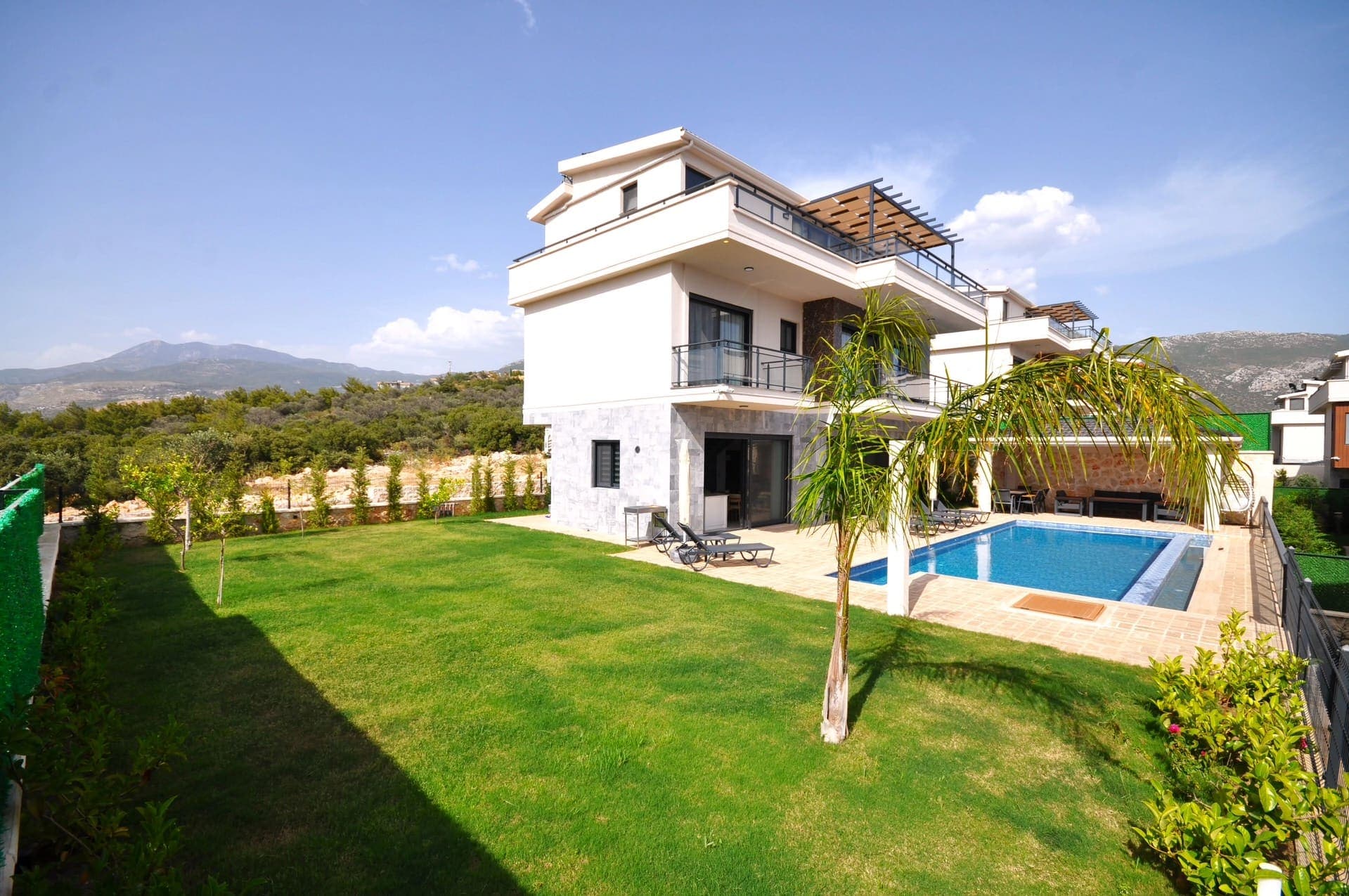 Yeşil bahçesi, özel yüzme havuzu ve oturma alanı bulunan iki katlı modern villa dış cephesi.