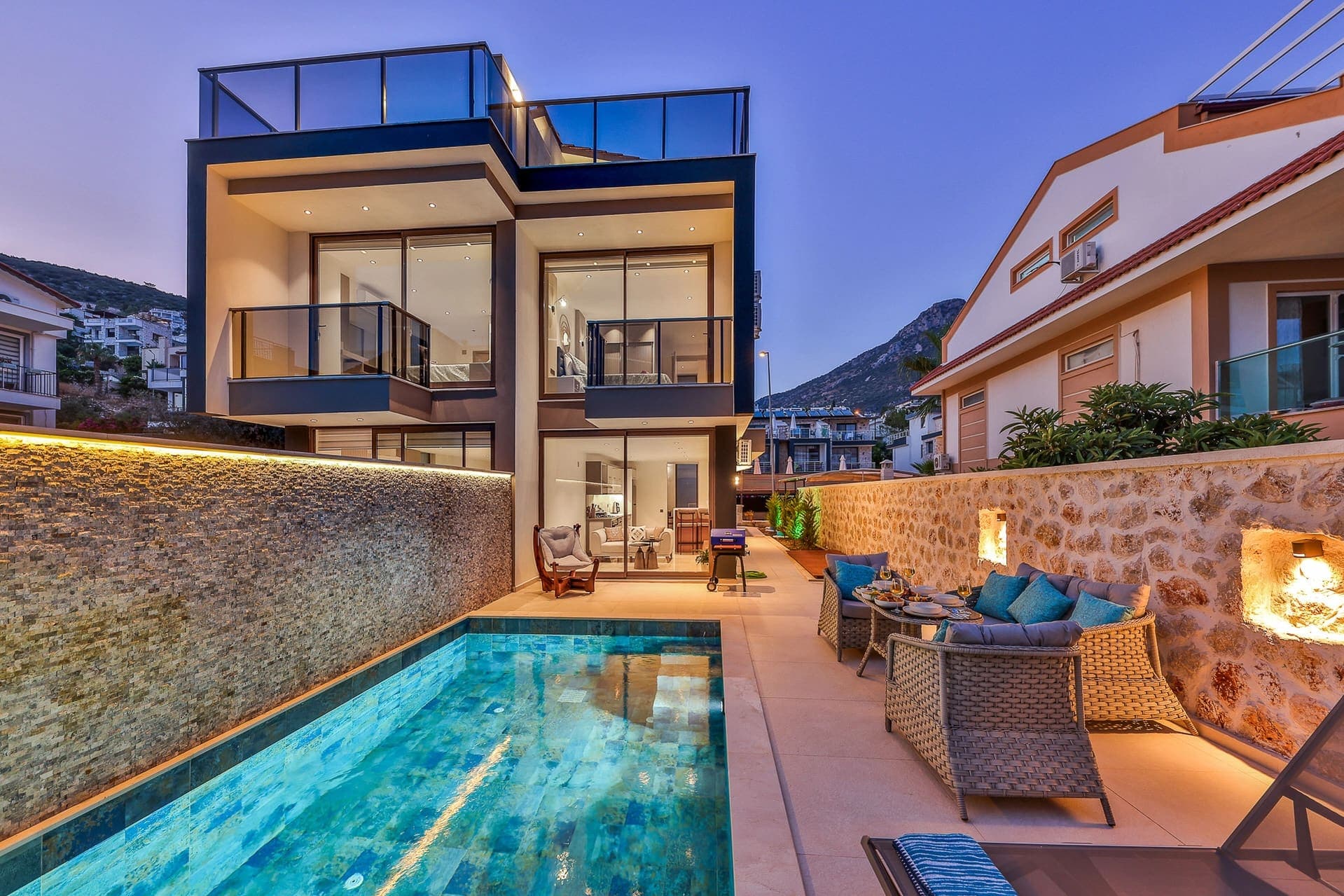 Akşam vakti aydınlatılmış modern villa, taş duvarlı özel havuz ve veranda mobilyaları.