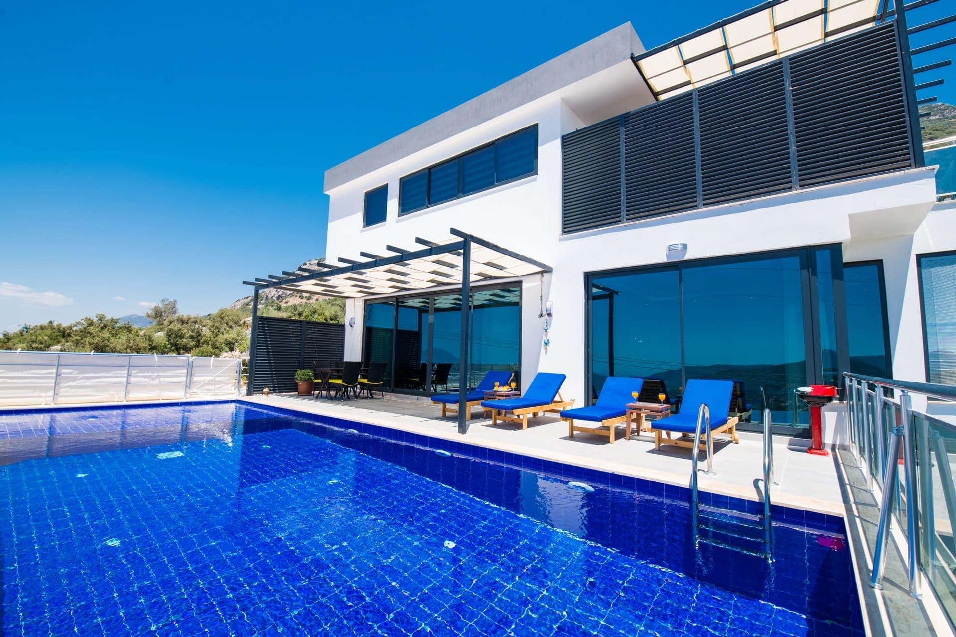 Güneşli havada, mavi havuz kenarında şezlonglar ve arka planda modern villa.