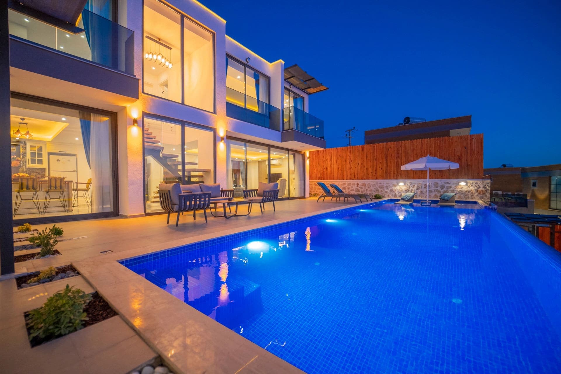 Gece aydınlatmasında havuzlu modern villa, terasta oturma grubu ve güneş şemsiyesi.