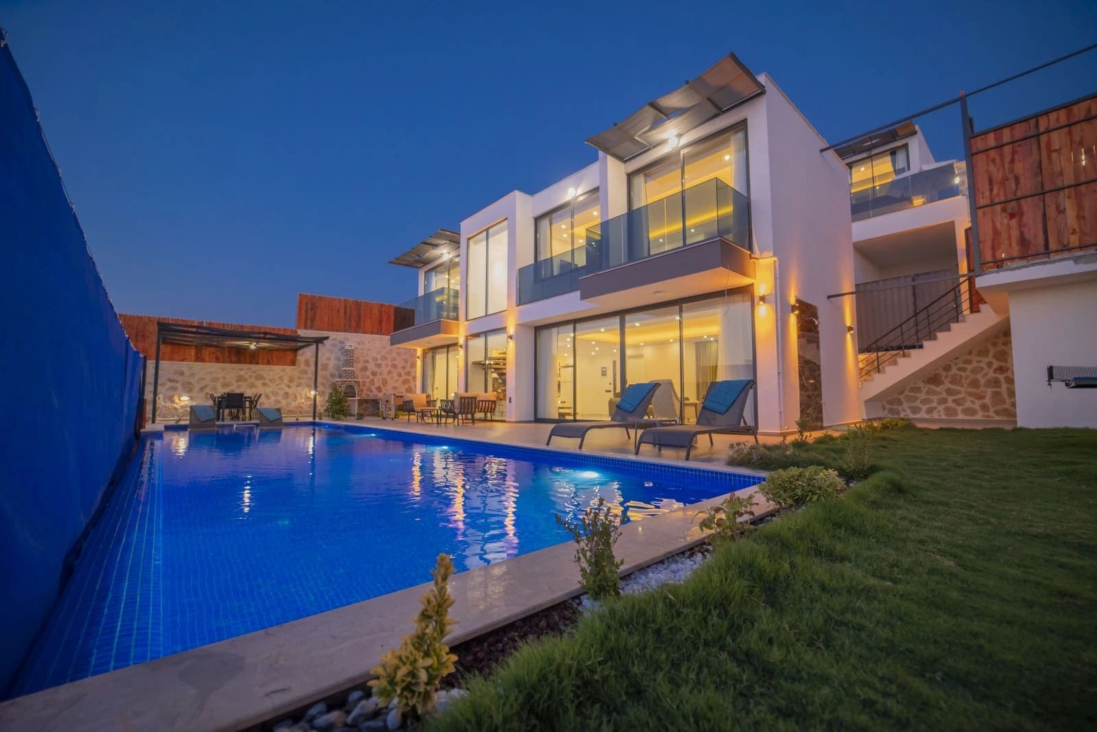 Gece ışıklandırılmış modern villa, önünde havuz, çim alan ve şezlonglar.