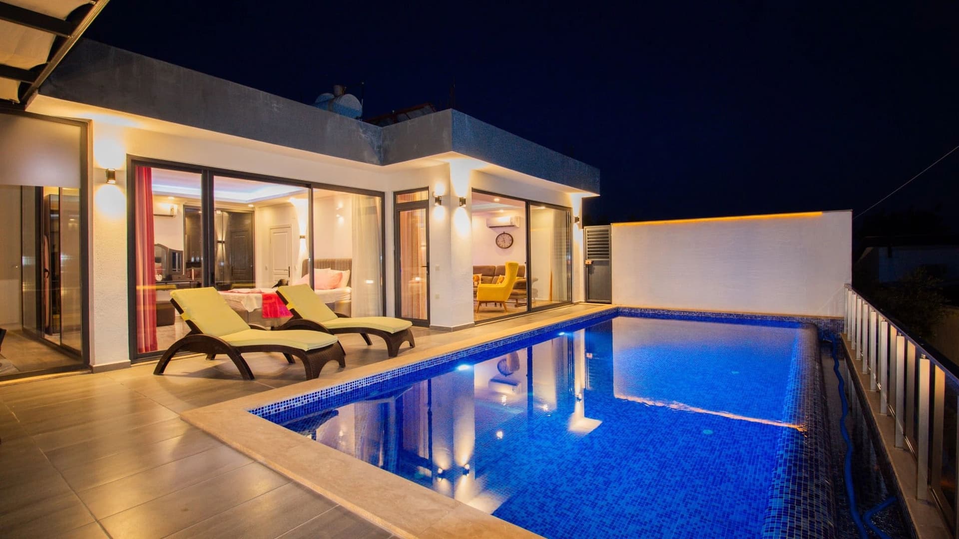 Gece aydınlatılmış havuz kenarında şezlonglar ve iç mekânı görünen modern villa.