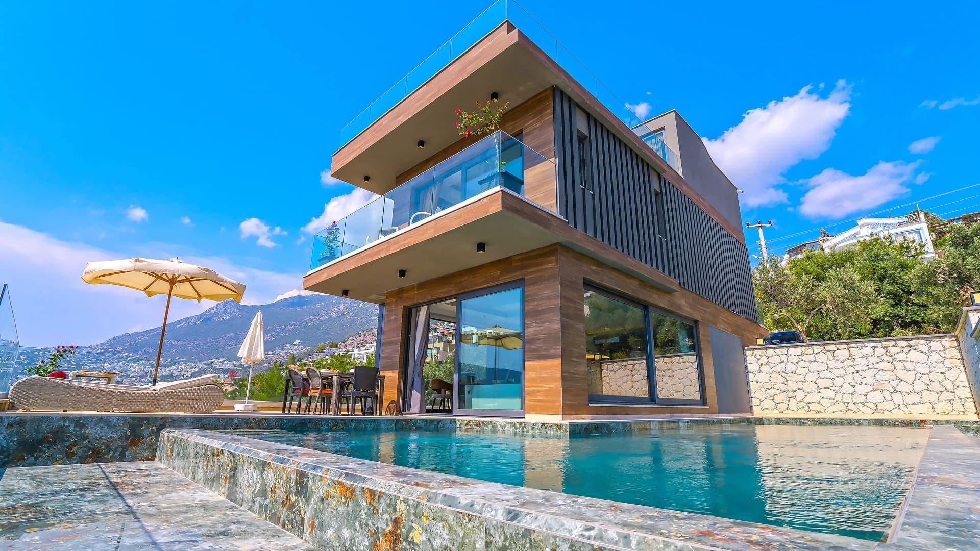 Ahşap kaplamalı, balkonlu, havuz başında şezlong ve yemek masası bulunan modern villa.