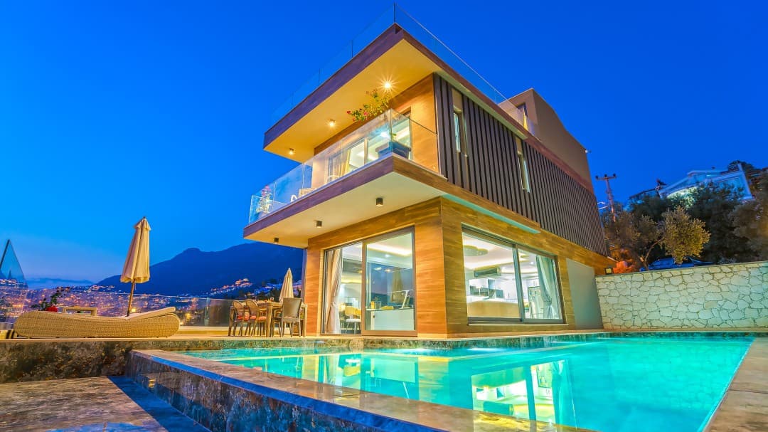 Akşam saatlerinde aydınlatılmış, havuzlu ve balkonlu modern villanın dış cephesi.