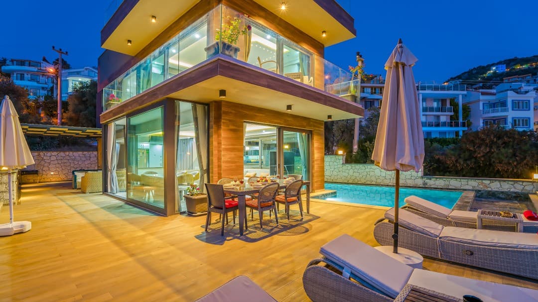 Akşam saatlerinde aydınlatılmış iki katlı modern villa ve havuzbaşı yemek masası.