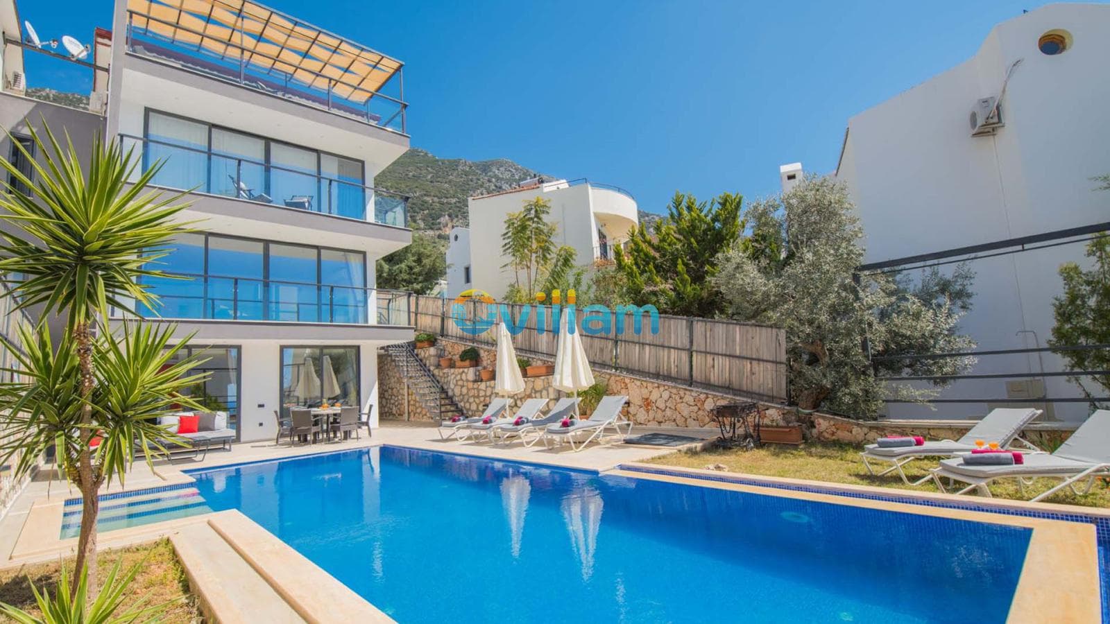 Çok katlı modern villa, önünde geniş yüzme havuzu, şezlonglar ve güneş şemsiyeleri.