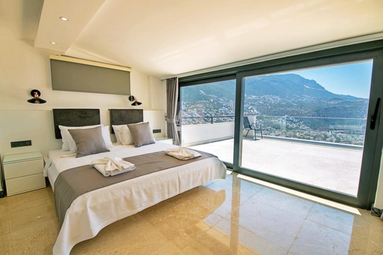 Çift kişilik yatak, bej mermer zemin ve panoramik deniz manzaralı geniş cam balkon kapısı.