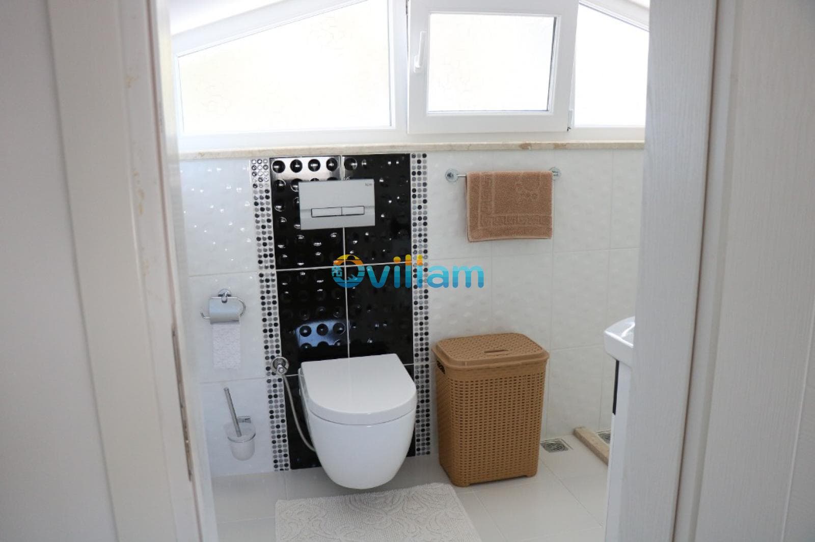 Beyaz fayanslı, asma klozetli ve hasır çamaşır sepeti bulunan küçük banyo.