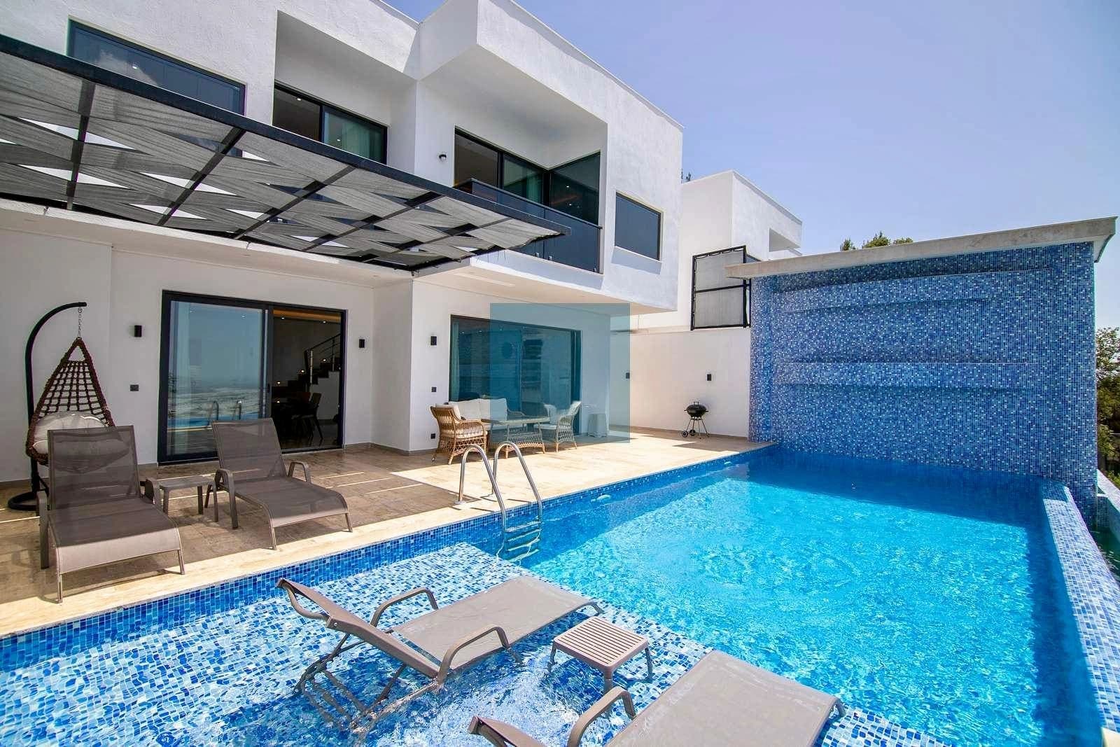Modern beyaz villa, önünde mavi yüzme havuzu, şezlonglar, asılı salıncak ve oturma alanı bulunuyor.
