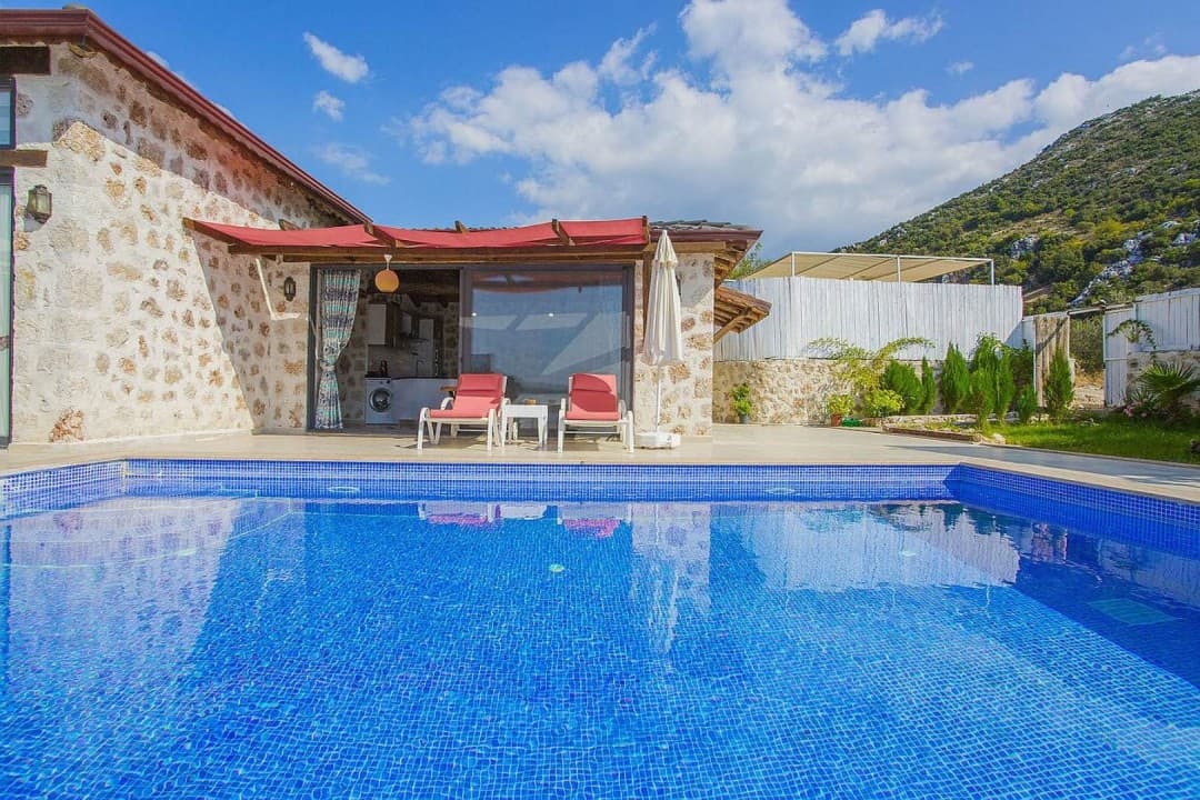 Mavi havuzun arkasında taş duvarlı villa, iki şezlong ve dağ manzarası.