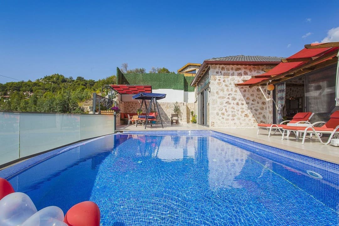 Mavi havuzun kenarında kırmızı minderli şezlonglar ve salıncak bulunan taş cepheli villa bahçesi.