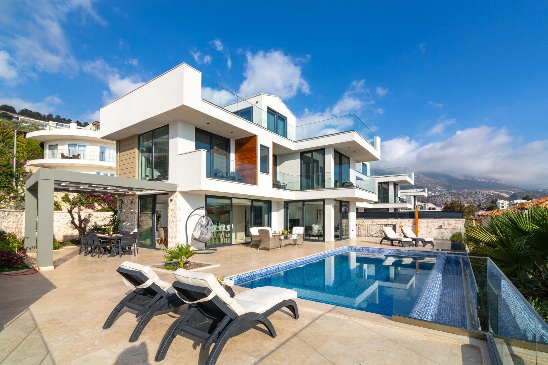 Modern beyaz villa, önünde havuz, şezlonglar ve yemek masası bulunan teras.