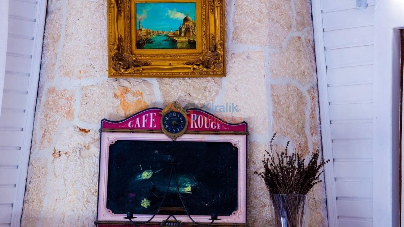 Taş duvar üzerinde asılı altın çerçeveli bir tablo ve altında Cafe Rouge yazılı dekoratif bir pano.