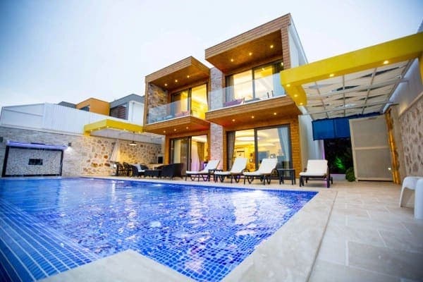 Ahşap detaylı modern iki katlı villa, önünde mavi mozaik kaplı yüzme havuzu ve şezlonglar yer alır.