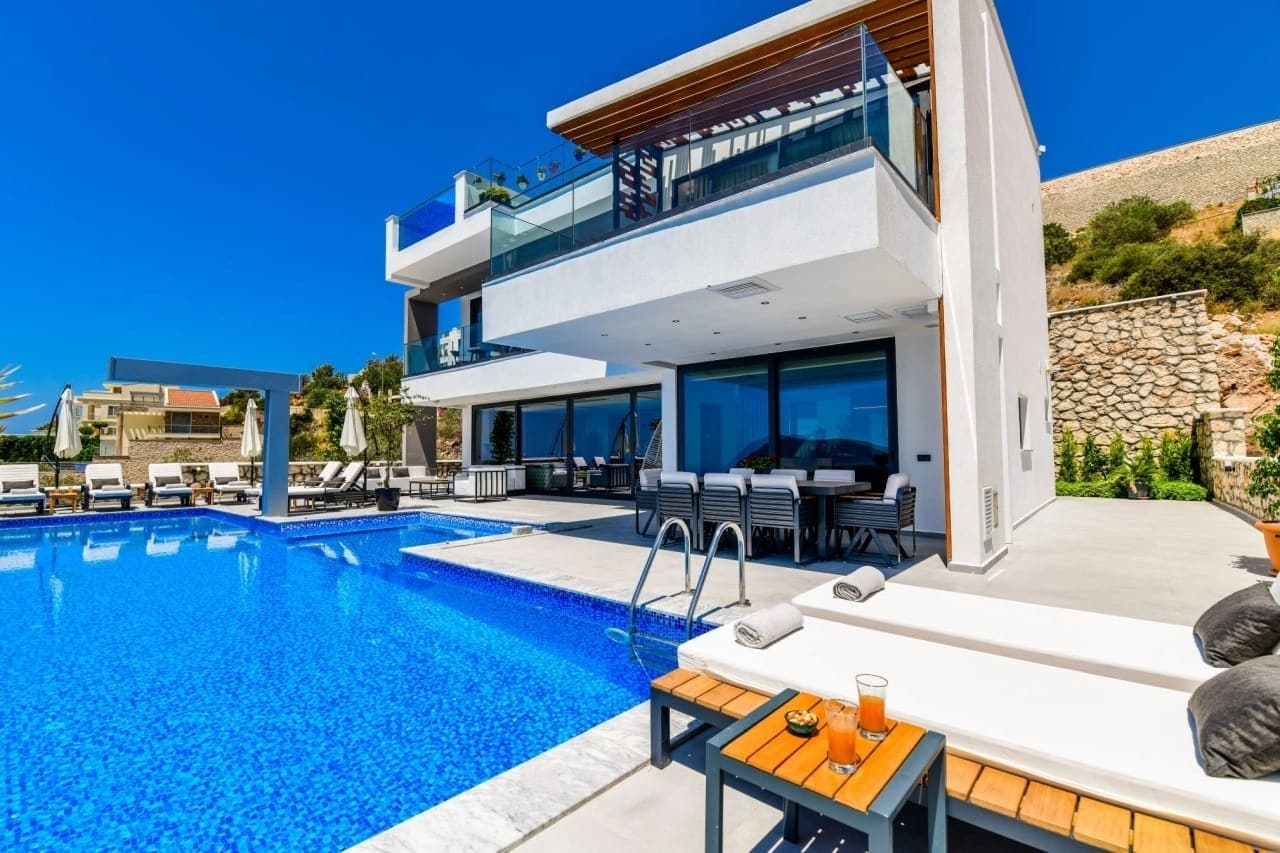 Beyaz dış cepheli modern villa, önünde mavi yüzme havuzu, şezlonglar ve yemek masası bulunur.