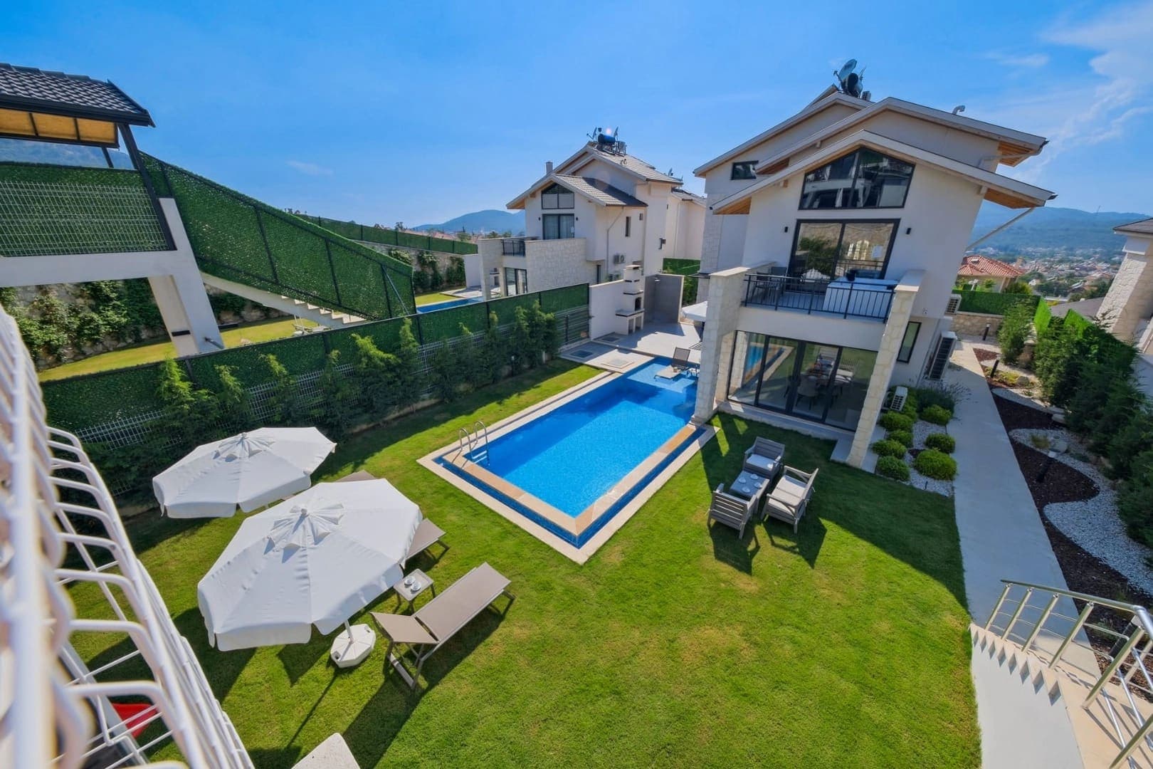 Bahçesinde havuz, şezlonglar ve beyaz şemsiyeler bulunan iki katlı müstakil villa.