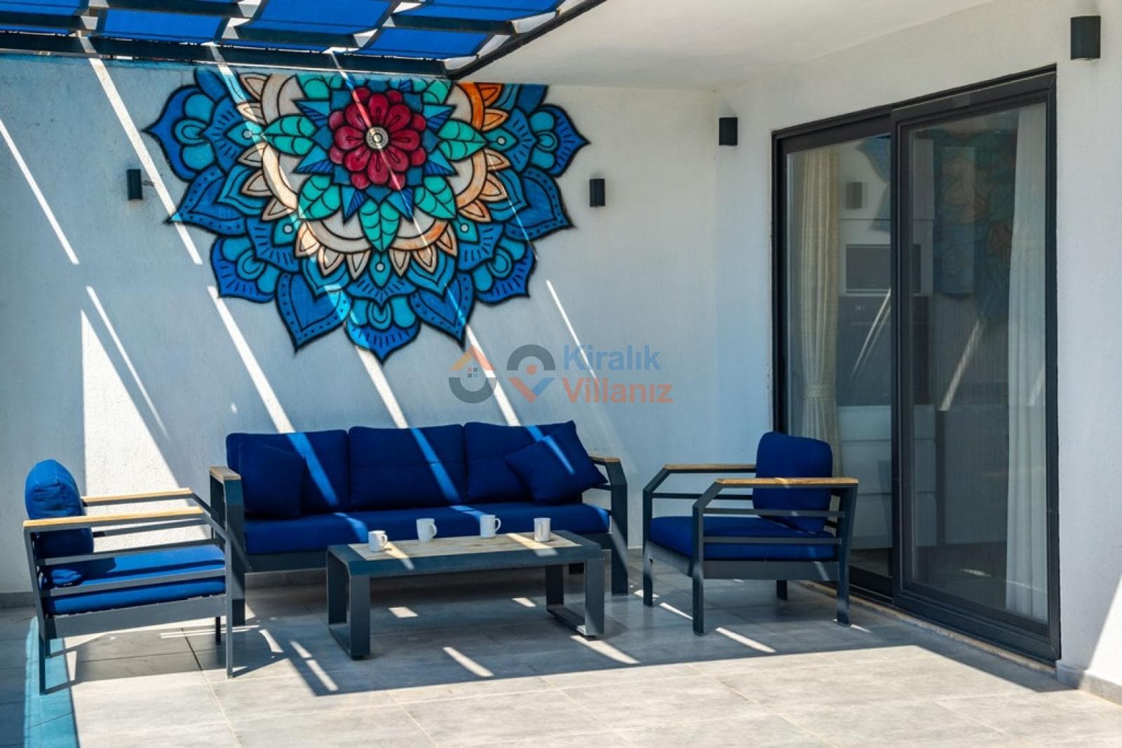 Duvarında renkli mandala deseni bulunan, mavi koltuk takımlı gölgelikli veranda.