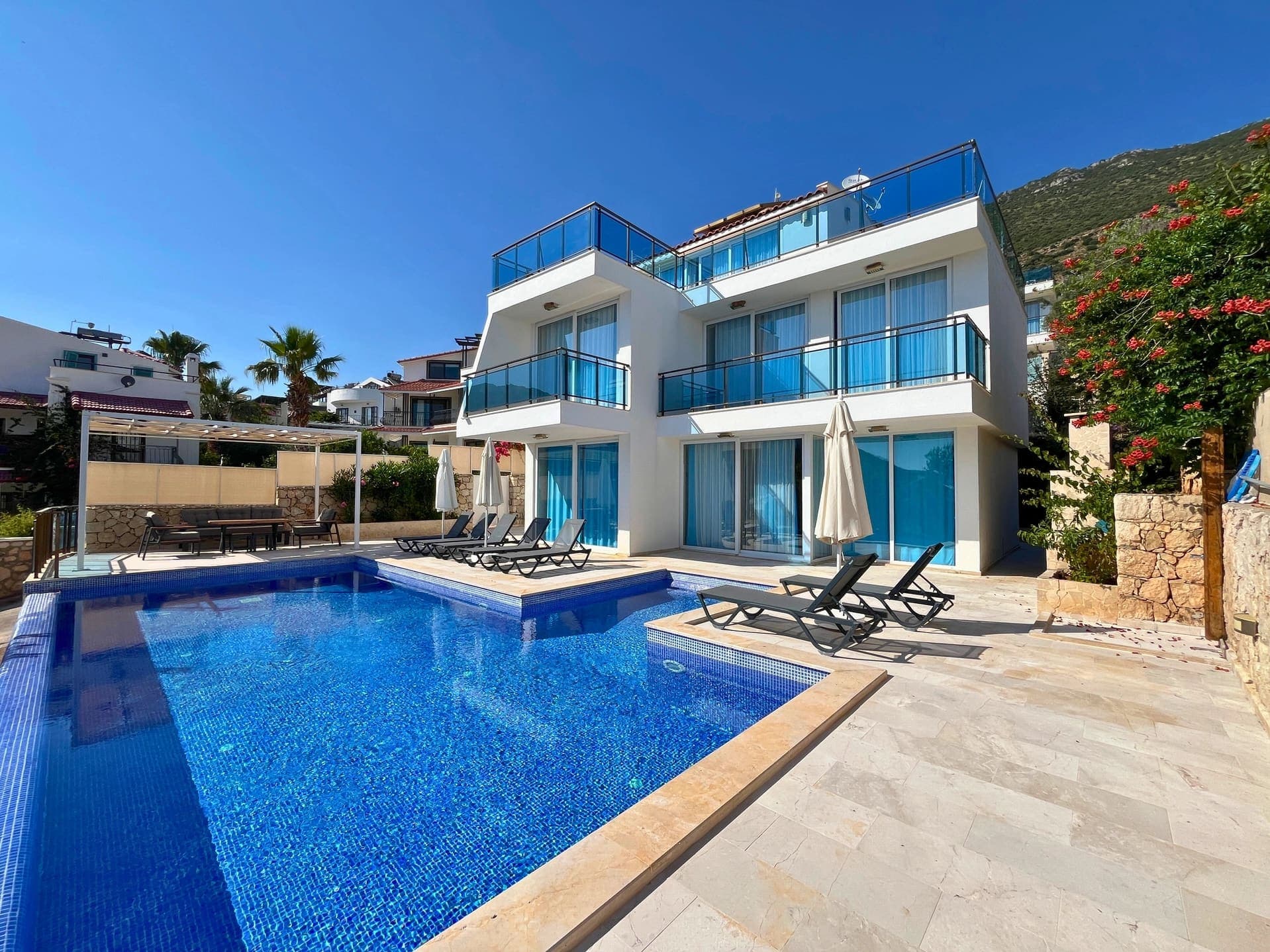 Mavi cam balkonlu, beyaz dış cepheli çok katlı villa ve önündeki güneşlenme teraslı havuz.