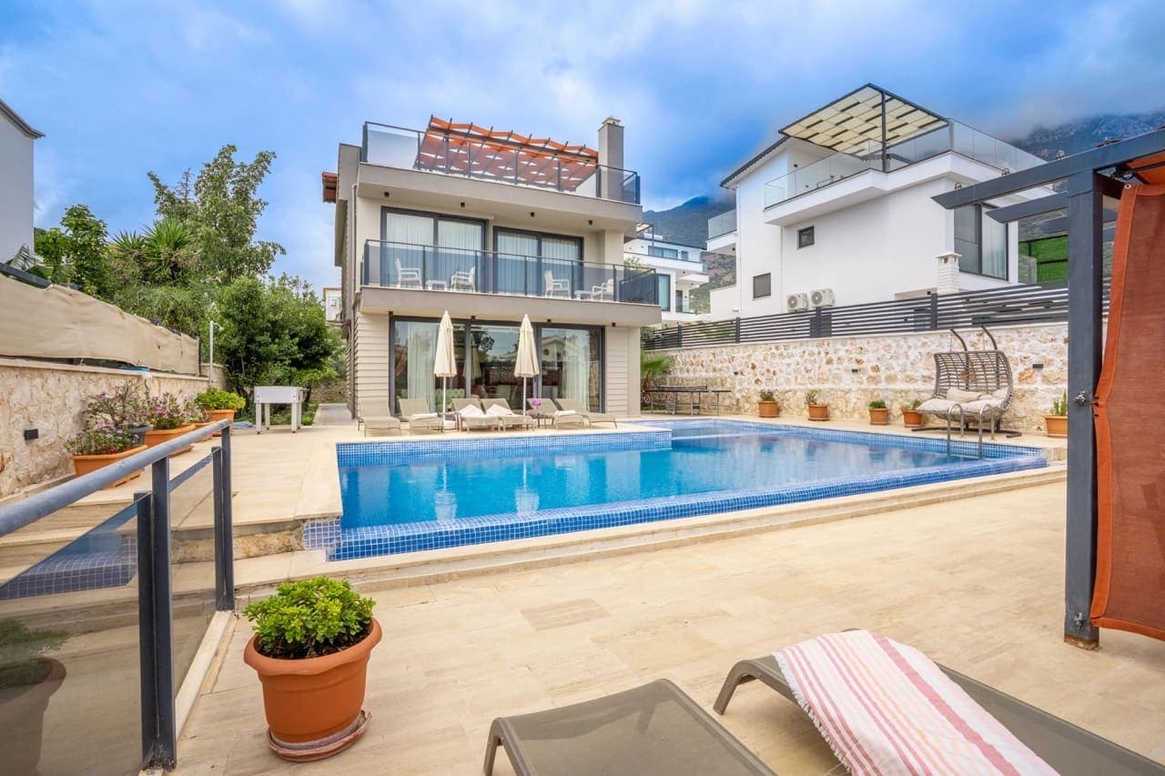 Modern iki katlı villa, önünde mavi yüzme havuzu, şezlonglar ve güneş şemsiyeleri ile çevrili teras.